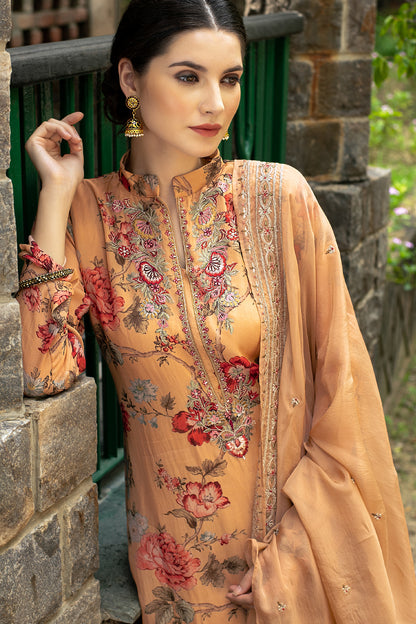 A woman models a peach-colored embroidered traditional outfit with floral patterns and a matching dupatta, set against a rustic stone wall and green fence background. | Wholesale Embroidered Muslin 3 Piece Kurta Set | Sri Fashion

