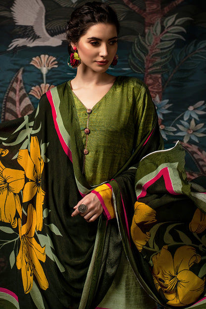 A woman models a three-piece olive green suit with a matching dupatta featuring a bold yellow floral print and pink and green borders. The background is a dark, ornate patterned tapestry. | Wholesale Pure Dola Silk Kurta Set For Women | Sri Fashion