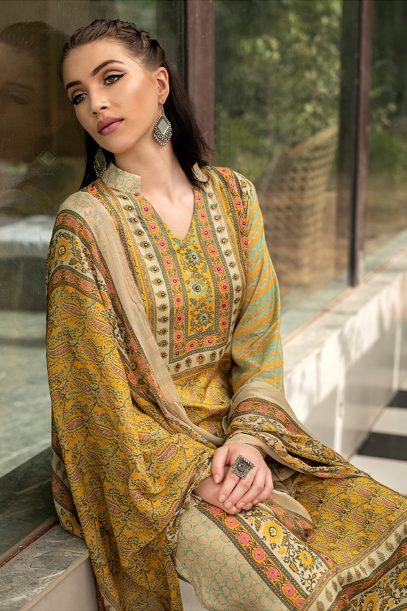 A woman seated indoors by a window wearing a patterned traditional outfit in mustard yellow, olive green, and beige with ornate border detailing. She is wearing large silver earrings and rings. | Wholesale Mustard Yellow Muslin Suit Set | Sri Fashion
