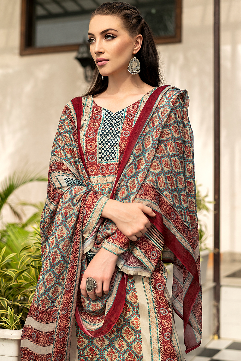 A woman models a traditional South Asian suit set featuring a printed suit and dupatta in red, beige, and teal, with a solid red border. She is wearing large silver earrings and standing in front of a neutral-toned building wall with some green plants visible. | Wholesale Off White Ethnic Muslin Suit Set | Sri Fashion

